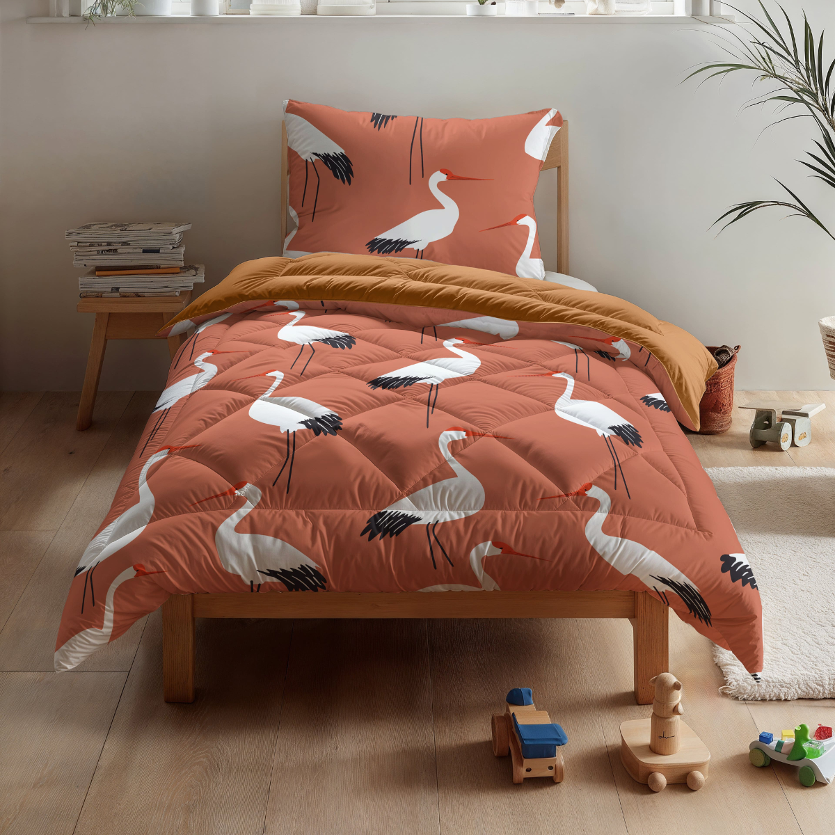 Das Zelesta Kiddybed Baumwolle - Terra Crane bietet orangefarbene Bettwäsche mit Kranich-Print, eine senfgelbe Decke und Kinderspielzeug aus Holz auf einem hellen Holzboden in einem hellen, minimalistischen Zimmer.