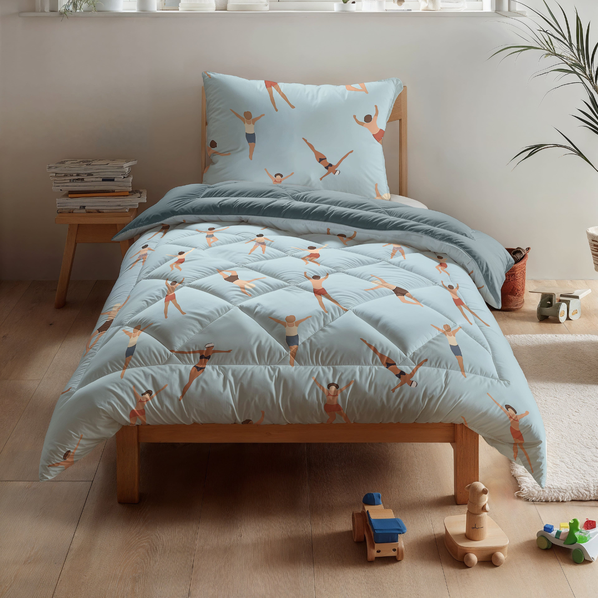 Ein Zelesta Kiddybed Baumwolle - Swimmers mit hypoallergenem Bettzeug und Schwimmeraufdrucken steht neben Holzspielzeug auf einem hellen Boden in einem hellen Zimmer.