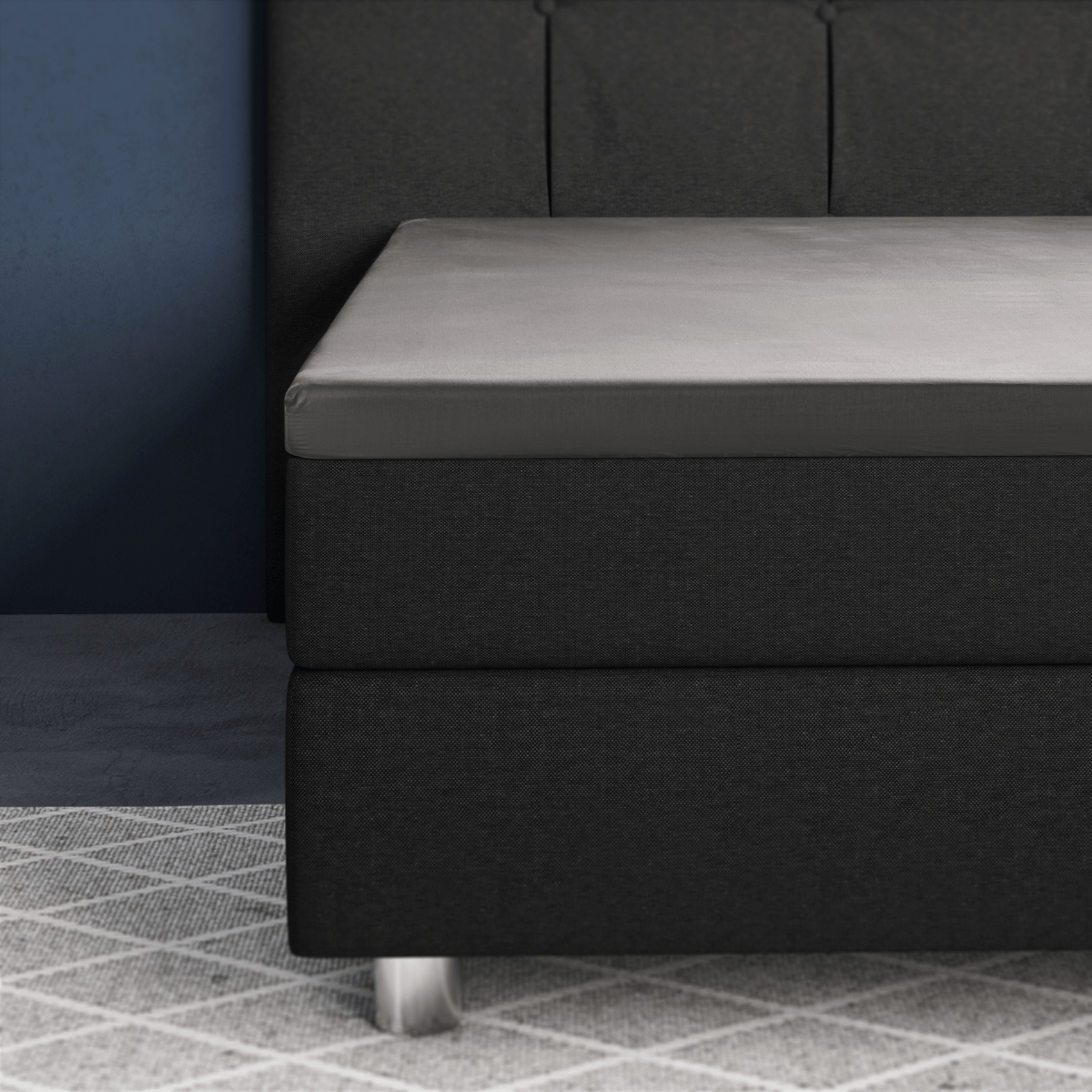 Nahaufnahme eines modernen Bettes mit dunklem Polsterrahmen, grauem Primaviera Deluxe Topper Satin Deluxe Spannbettuch und Metallfüßen auf einem hell gemusterten Teppich neben einer blauen Wand.