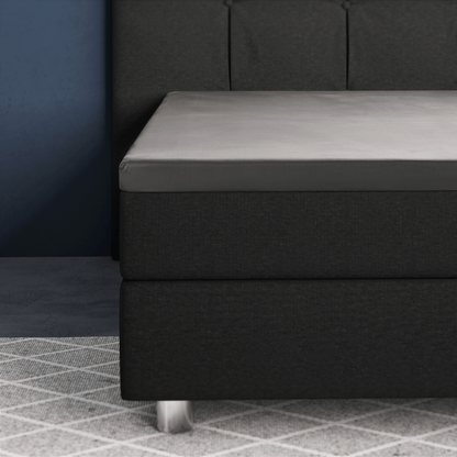 Nahaufnahme eines modernen Bettes mit dunklem Polsterrahmen, grauem Primaviera Deluxe Topper Satin Deluxe Spannbettuch und Metallfüßen auf einem hell gemusterten Teppich neben einer blauen Wand.