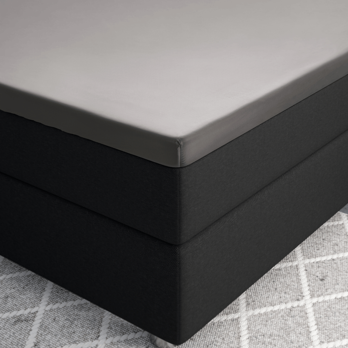 Nahaufnahme einer Bettecke mit dem Primaviera Deluxe Topper Satin Deluxe Spannbettuch in Grau auf einem dunklen Sockel, der auf einem hellgrau gemusterten Teppich liegt.