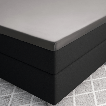 Nahaufnahme einer Bettecke mit dem Primaviera Deluxe Topper Satin Deluxe Spannbettuch in Grau auf einem dunklen Sockel, der auf einem hellgrau gemusterten Teppich liegt.