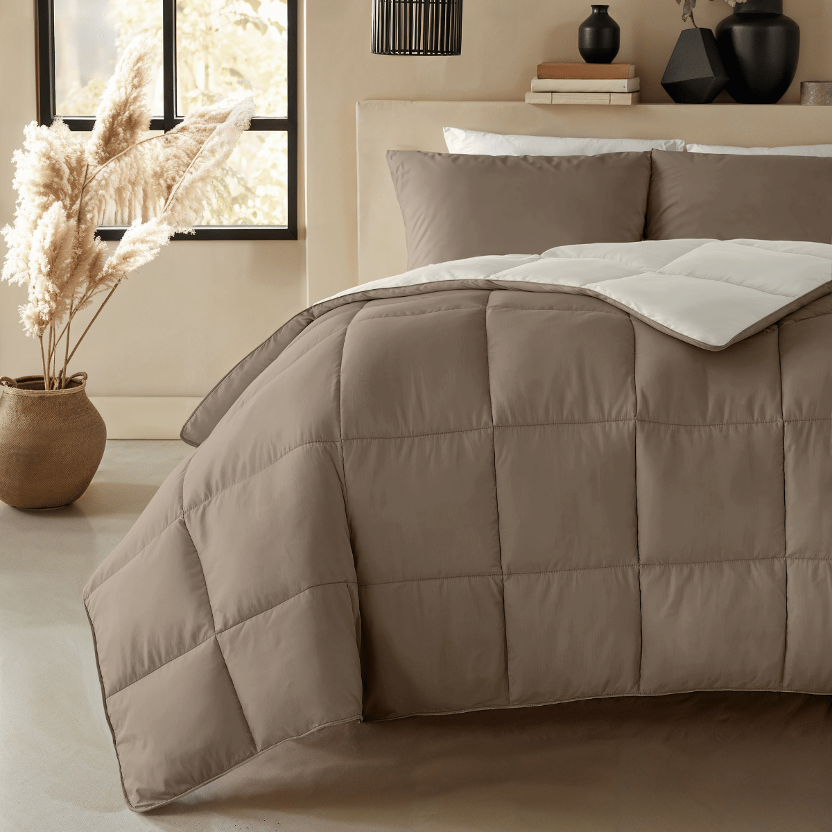 Ein ordentlich gemachtes Bett mit der Zelesta Premiumbed Baumwolle - Taupe & Cream Steppdecke, weißen Kissen und beiger Dekoration mit einer Pampasgrasvase neben dem Fenster.
