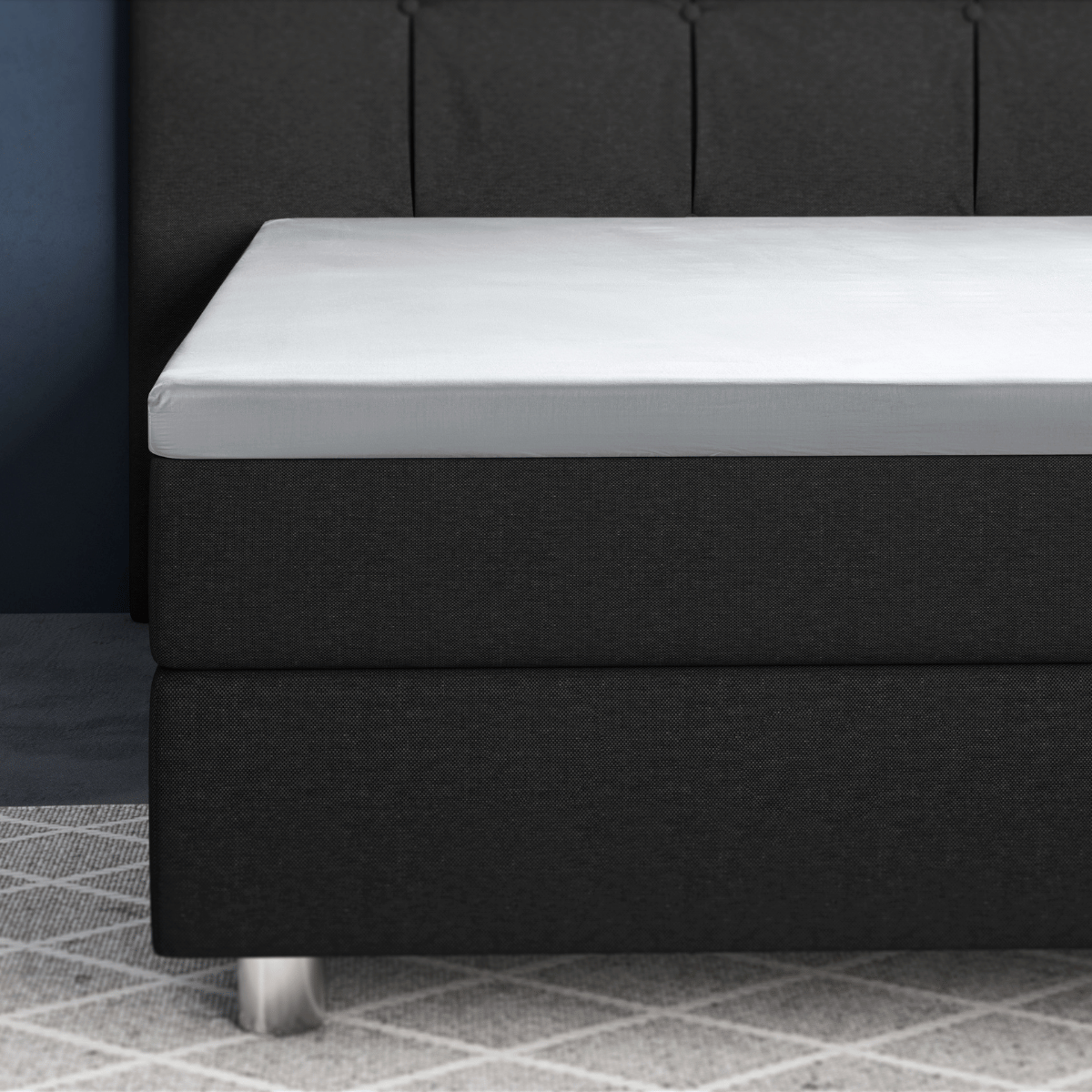 Nahaufnahme eines modernen Bettes mit dunklem Lattenrost und dem Primaviera Deluxe Topper Satin Deluxe Spannbettuch in Weiss über der Matratzenauflage, auf einem hell gemusterten Teppich.