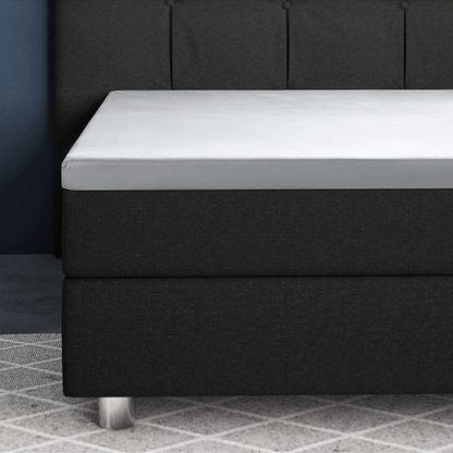 Nahaufnahme eines modernen Bettes mit dunklem Lattenrost und dem Primaviera Deluxe Topper Satin Deluxe Spannbettuch in Weiss über der Matratzenauflage, auf einem hell gemusterten Teppich.
