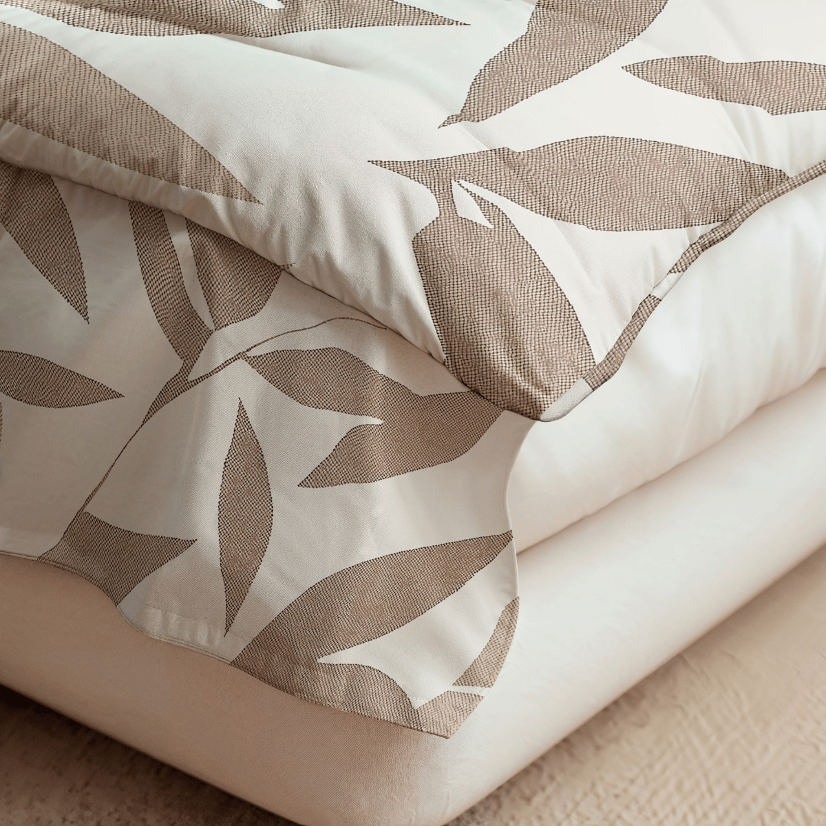 Nahaufnahme einer ordentlich gefalteten Royalbed Baumwolle - Beige Autumn Bettdecke mit einem beige-weißen Blattmuster auf einem schlichten weißen Laken - ideal als allergikergeeignete Bettdecke.