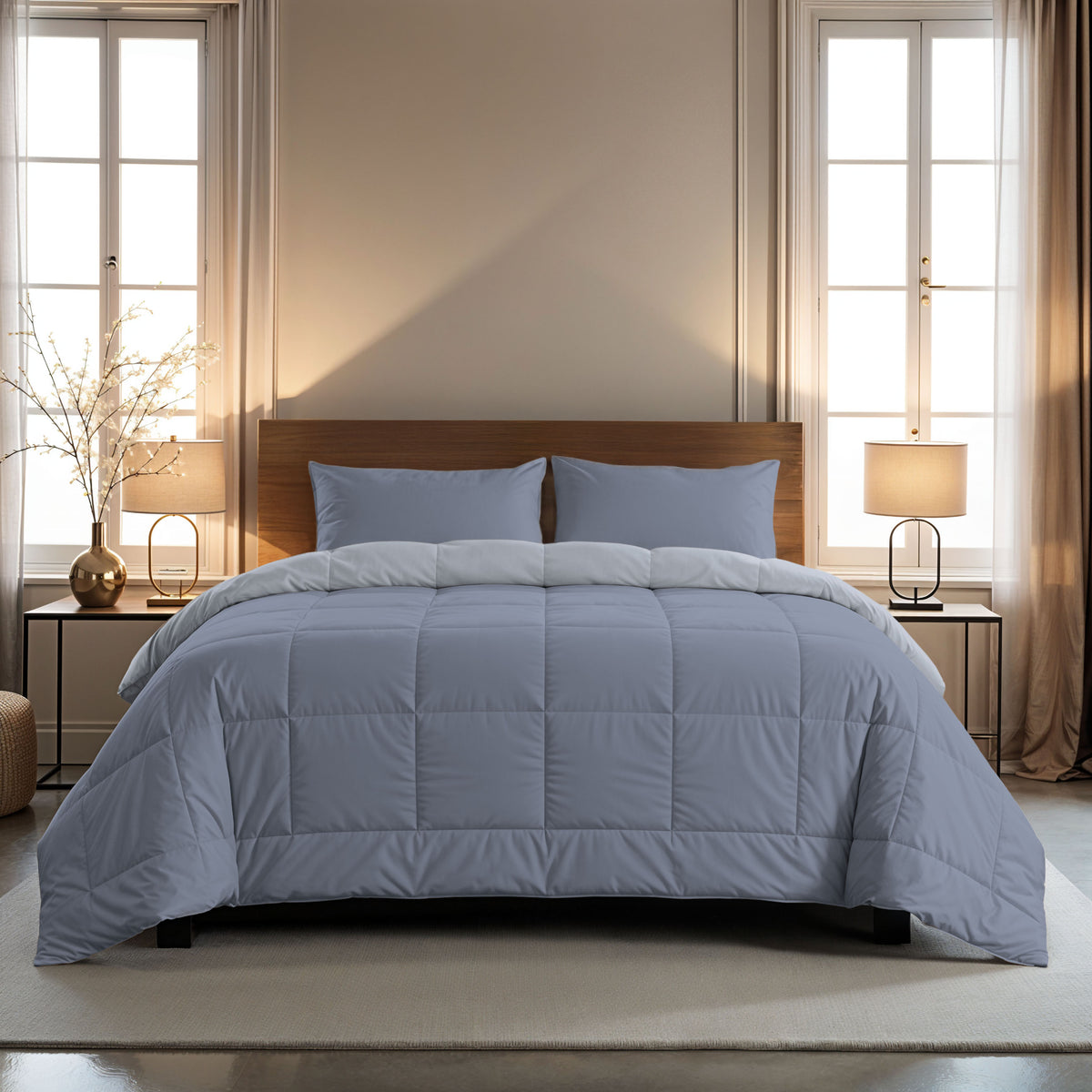 Ein ordentlich gemachtes Bett mit einer Zelesta Comfort Duo Bettdecke in Eisblau & Sanftes Blau und zwei Kissen, in der Mitte zwischen Fenstern mit beigen Vorhängen, Beistelltischen und Lampen in einem modernen Schlafzimmer.