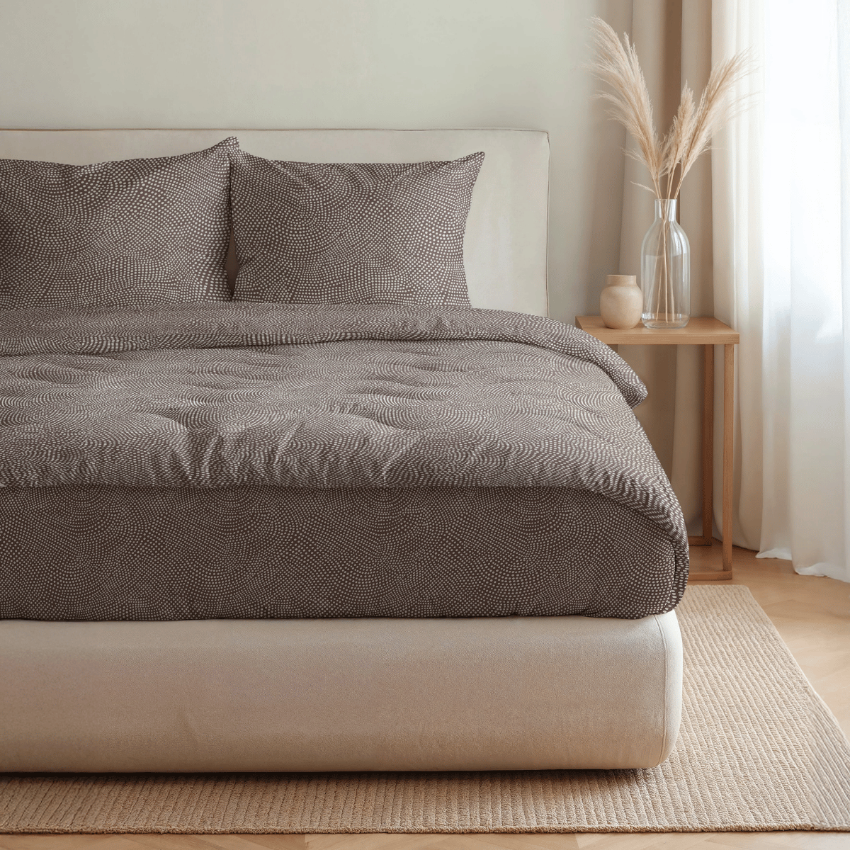 Ein ordentlich gemachtes Bett mit gemusterter brauner Bettwäsche und zwei Kissen, mit der Royalbed Baumwolle - Brown Animal Bettdecke für Allergiker, steht neben einem hölzernen Nachttisch mit einer Vase mit getrockneten Gräsern und einem Dekorationsartikel.