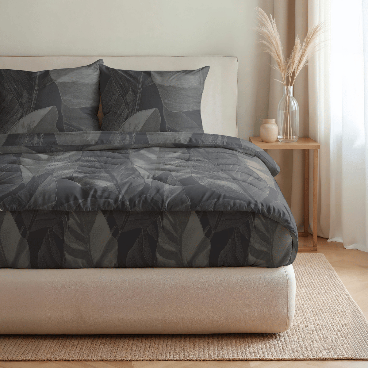 Ein ordentlich gemachtes Bett mit der Royalbed Baumwolle - Black Swan blattgemusterten Bettwäsche und Kissen, plus einer temperaturregulierenden Bettdecke, sitzt neben einem hölzernen Nachttisch mit getrocknetem Pampasgras in einem sanft beleuchteten Raum.