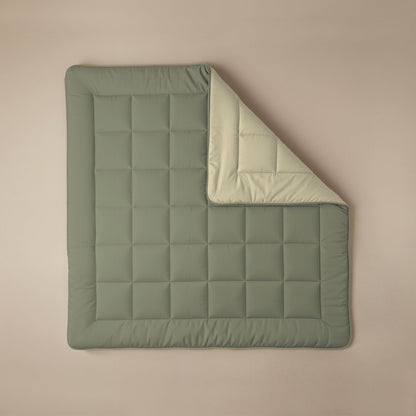Die Zelesta Comfort Duo Bettdecke in olivgrün & creme liegt flach auf einer beigen Fläche mit abgerundeten Kanten, wobei eine Ecke zurückgeklappt ist und die hellere cremefarbene Unterseite zeigt.