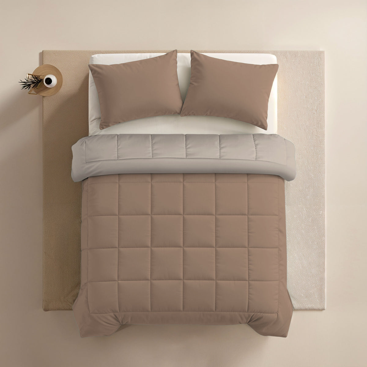 Ein ordentlich gemachtes Bett mit hellbraunen Kissen, einer Zelesta Comfort Duo Bettdecke - Terra & Sand, einem hellgrauen Laken und einem kleinen runden Nachttisch mit einer Vase, die einen einzelnen Zweig enthält.