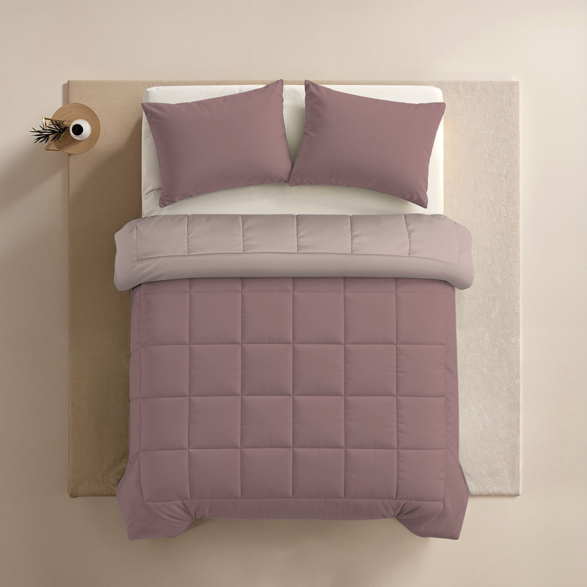 Ein gemachtes Bett mit mauvefarbenen Kissen in 80x80cm Zelesta Comfort Duo Kissenbezüge Set von 2 - Rosa, eine passende gesteppte Bettdecke, ein hellbeiges Oberlaken und ein kleiner runder Beistelltisch mit einer Vase in der oberen linken Ecke.