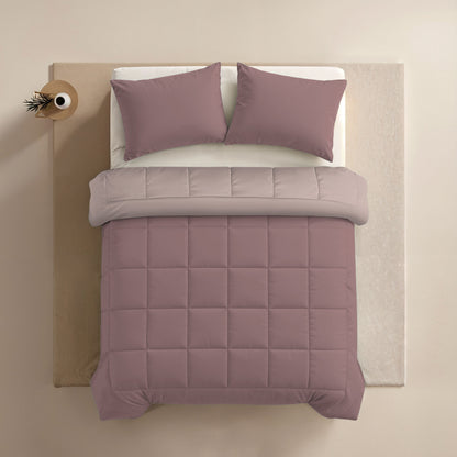 Ein gemachtes Bett mit mauvefarbenen Kissen in 80x80cm Zelesta Comfort Duo Kissenbezüge Set von 2 - Rosa, eine passende gesteppte Bettdecke, ein hellbeiges Oberlaken und ein kleiner runder Beistelltisch mit einer Vase in der oberen linken Ecke.