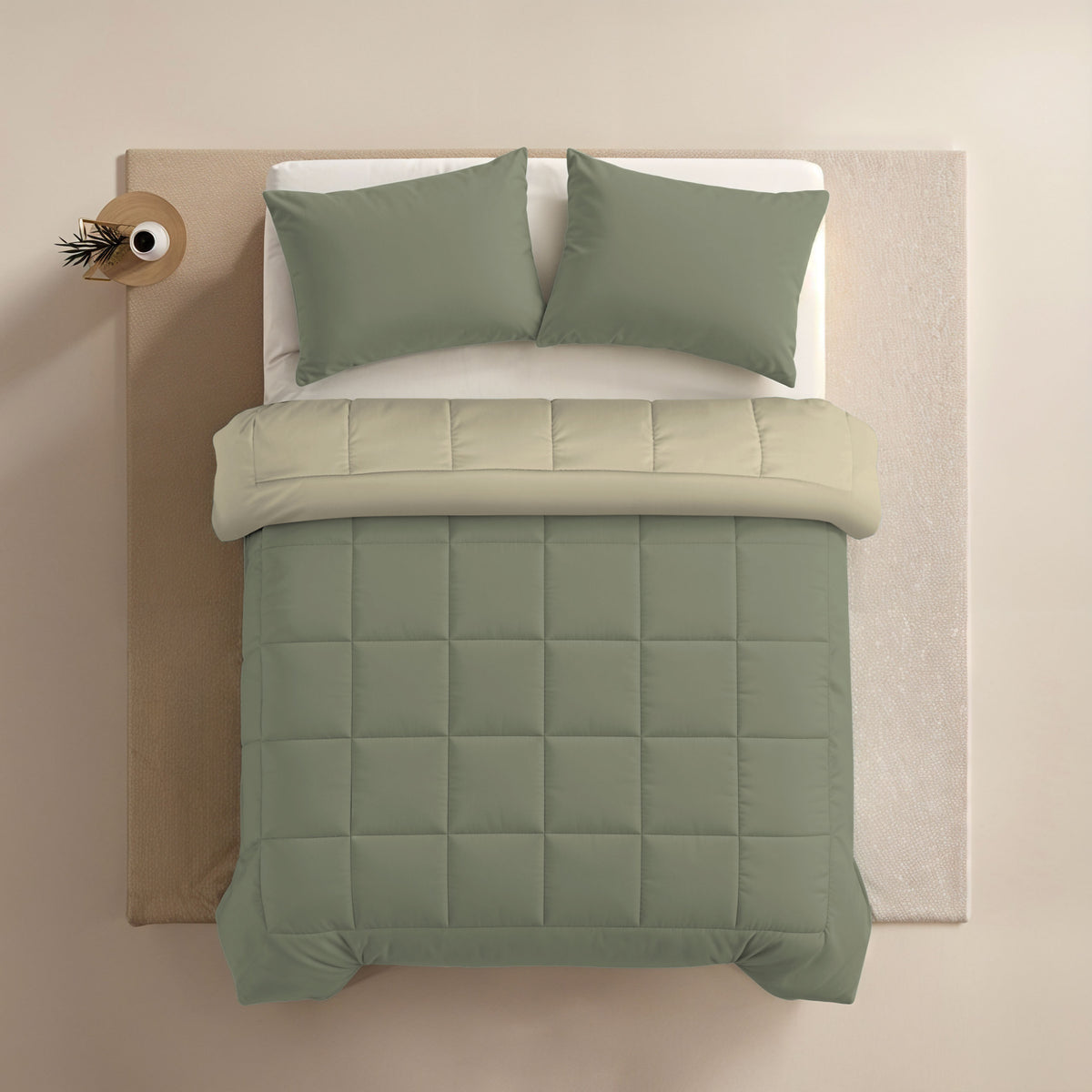 Blick von oben auf ein ordentlich gemachtes Bett mit salbeigrüner Bettwäsche, zwei passenden Kissen und einem kleinen runden Nachttisch mit einer Pflanze. Die Einrichtung besteht aus der Zelesta Comfort Duo Bettdecke - Olivgrün & Creme für ganzjährigen Komfort.