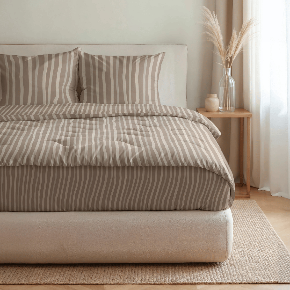 Ein ordentlich gemachtes Bett mit der Royalbed Baumwolle - Brown Stripes Bettdecke, zwei Kissen, ein hölzerner Nachttisch mit einer Vase mit getrocknetem Gras und einer Kerze, und ein allergikergeeigneter heller Teppich auf einem Holzboden.