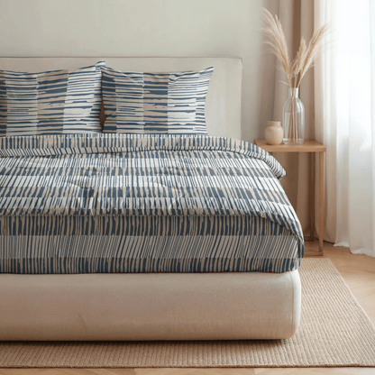 Auf einem ordentlich gemachten Bett mit blau-weiß gestreifter Bettwäsche, zwei passenden Kissen und einem Nachttisch mit Vasen mit getrockneten Gräsern liegt die Royalbed Baumwolle - Checkerboard, eine temperaturregulierende und allergikerfreundliche Bettdecke.