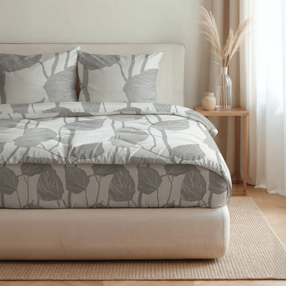 Ein ordentlich gemachtes Bett mit Royalbed Baumwolle - Spring Buds grauer, blattgemusterter Bettwäsche und einer temperaturregulierenden Bettdecke steht neben einem hölzernen Beistelltisch mit einer Glasvase, Pampasgras, Kerze und Keramiktasse in einem sanft beleuchteten Schlafzimmer.