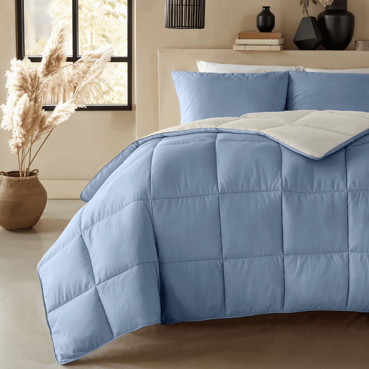 Ein Zelesta Premiumbed Baumwolle - Dream Blue & Cream mit passenden Kissen steht in einem beigefarbenen Raum, der mit getopften Pampasgräsern geschmückt und in natürliches Licht getaucht ist.
