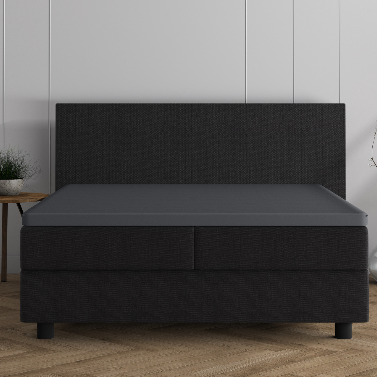 Ein minimalistisches Schlafzimmer mit einem schwarzen Polsterbett mit hohem Kopfteil, bezogen mit einem Doppel Jersey Topper Spannbettlaken - Anthrazit, vor einer weiß getäfelten Wand und einem Holzboden. Auf der linken Seite befindet sich ein kleiner Beistelltisch mit einer Pflanze.
