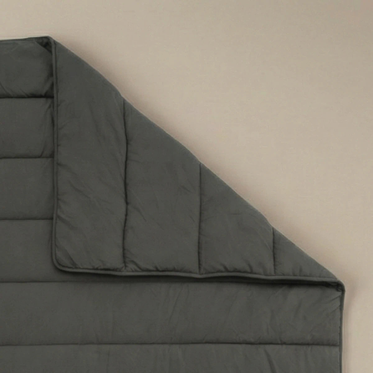 Eine Zelesta Comfort Uni Bettdecke in Anthrazit liegt flach auf einer beigen Oberfläche, wobei eine Ecke umgeschlagen ist, um die detaillierten Nähte und die weiche Textur zu zeigen.