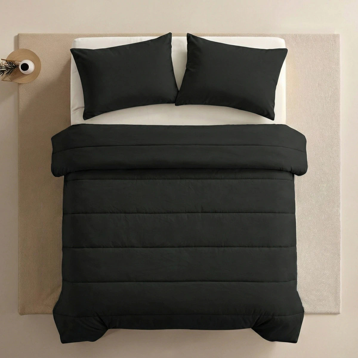 Ein ordentlich gemachtes Bett mit einer schwarzen Bettdecke und zwei Zelesta Comfort Uni Kissenbezügen Set von 2 - Schwarz (80x80cm), platziert auf einem hellen Teppich neben einem kleinen runden Beistelltisch mit einer Vase.