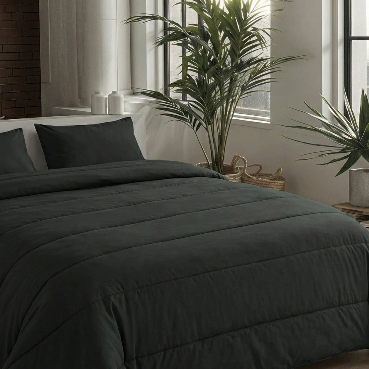 Ein ordentlich gemachtes Bett mit zwei Zelesta Comfort Uni Kissenbezügen 80x80cm in Schwarz, positioniert durch große Fenster und hohe Grünpflanzen in einem modernen, minimalistischen Schlafzimmer.