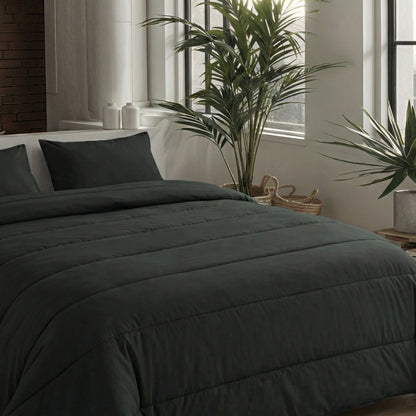 Ein ordentlich gemachtes Bett mit zwei Zelesta Comfort Uni Kissenbezügen 80x80cm in Schwarz, positioniert durch große Fenster und hohe Grünpflanzen in einem modernen, minimalistischen Schlafzimmer.