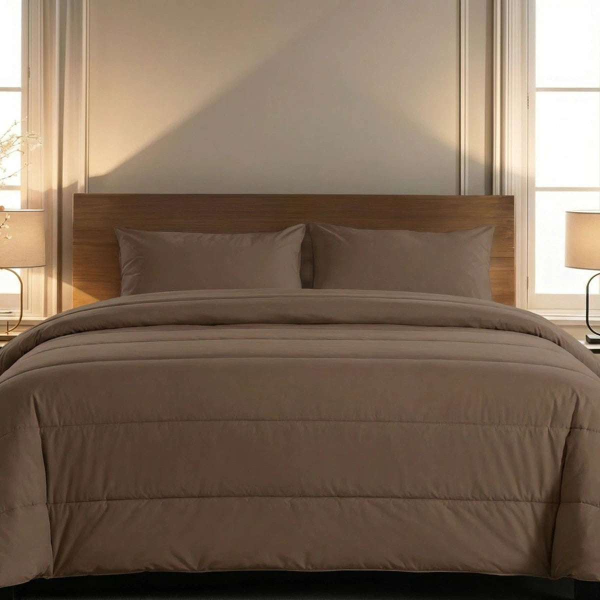 Ein ordentlich gemachtes Bett mit dem Zelesta Comfort Uni Kissenbezüge Set von 2 in Mokka (80x80cm), zwei Kissen, einem Kopfteil aus Holz, Nachttischlampen und ist an einer Wand mit zwei großen Fenstern aufgestellt.