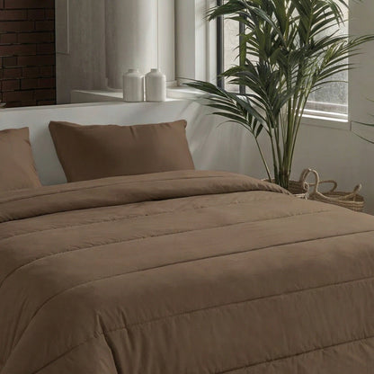 Ein hübsch gemachtes Bett mit braunen Zelesta Comfort Uni Kissenbezügen Set von 2 - Mokka (80x80cm) und einer Bettdecke steht neben einer großen Grünpflanze und einem Fenster. Zwei weiße Deko-Gläser stehen auf einem Regal hinter dem Bett.
