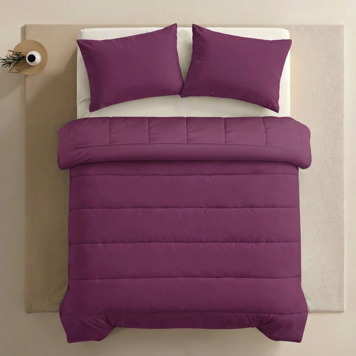 Ein ordentlich gemachtes Bett mit einer lila Bettdecke und zwei passenden 80x80cm Zelesta Comfort Uni Kissenbezügen Set von 2 in Bordeauxrot, platziert auf einem beigen Podest vor einem hellen Hintergrund.