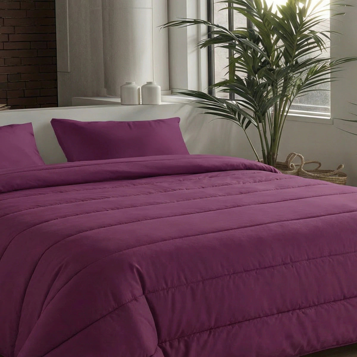 Das Zelesta Comfort Uni Kissenbezüge Set von 2 in Bordeauxrot (80x80cm), gepaart mit einer passenden Mikrofaser-Bettdecke, stehen in einem ordentlich gemachten Bett neben einem Fenster mit einer Topfpflanze und weißem Dekor.