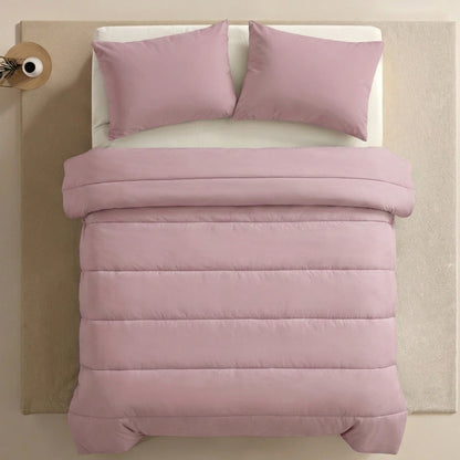 Ein ordentlich gemachtes Bett mit der Zelesta Comfort Uni Bettdecke in altrosa und zwei passenden Kissen ruht auf einem beigen Teppich neben einem kleinen runden Nachttisch mit einer Lampe.