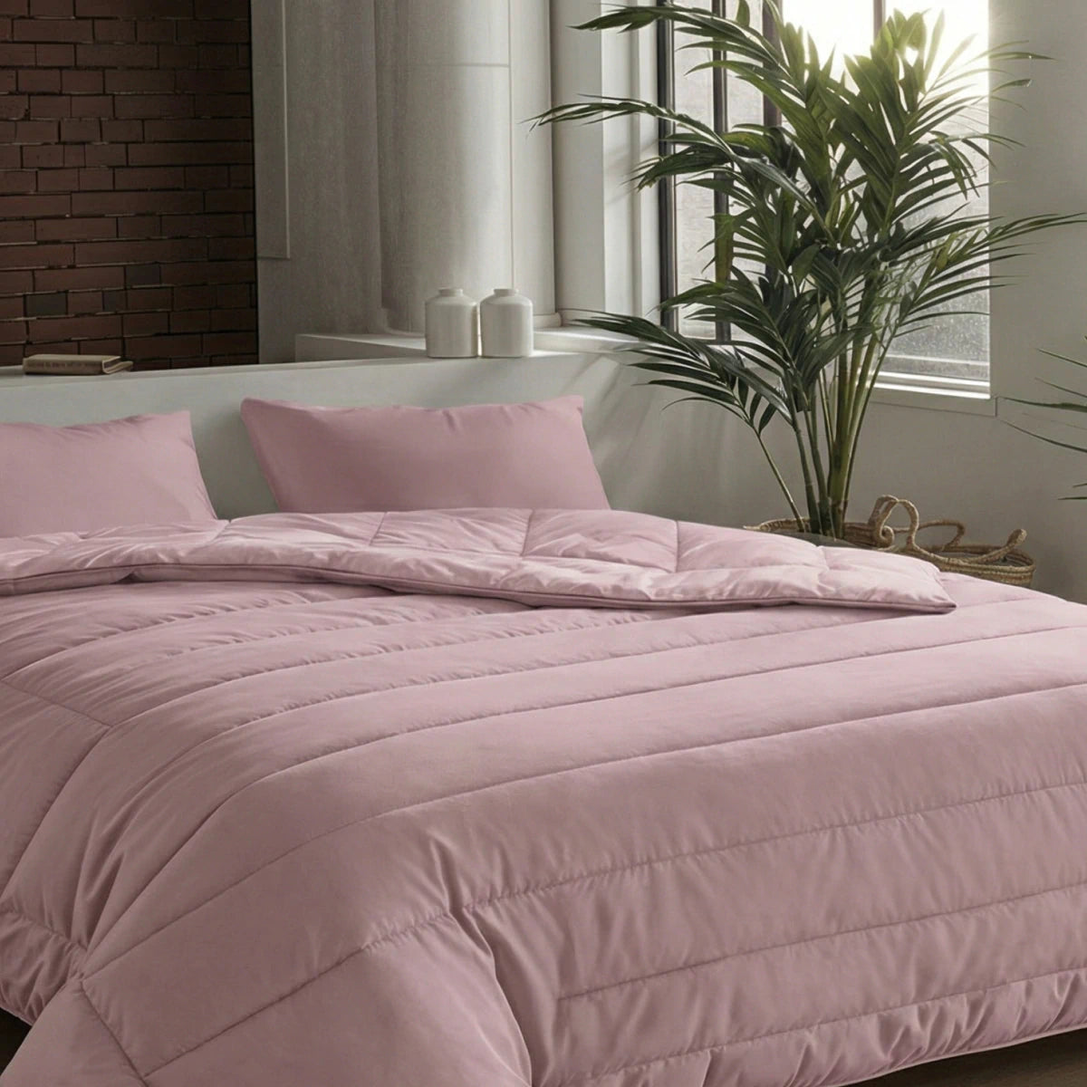 Ein ordentlich gemachtes Bett mit einer Zelesta Comfort Uni Bettdecke in altrosa und passenden Kissen steht in einem modernen Schlafzimmer mit großen Fenstern, einer Backsteinwand und einer hohen Grünpflanze.