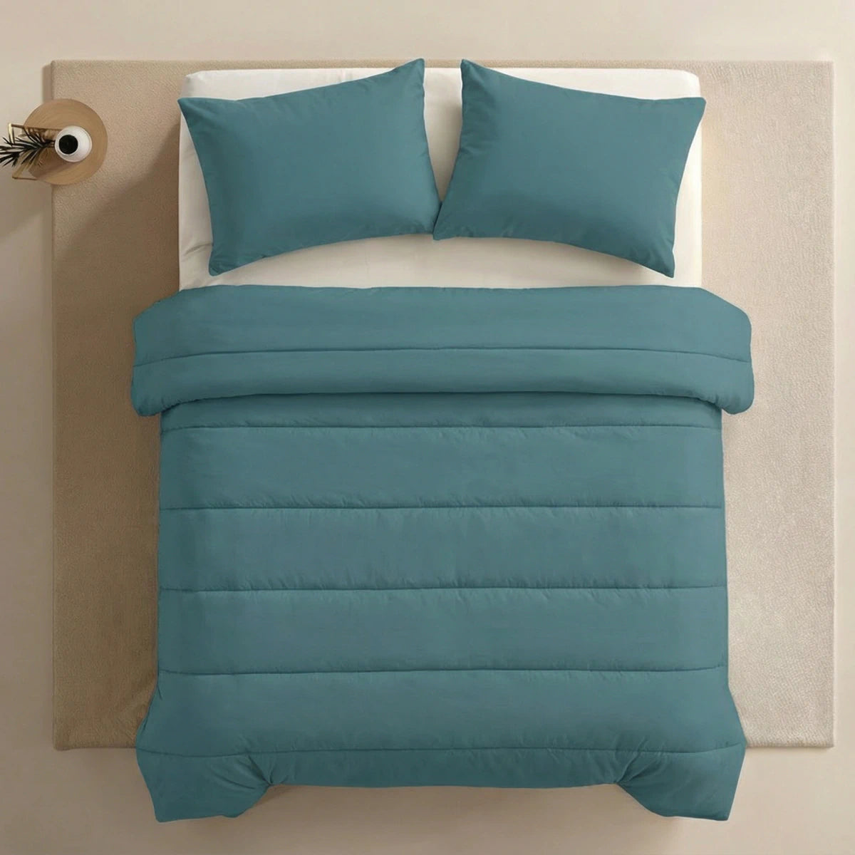 Ein ordentlich gemachtes Bett mit Zelesta Comfort Uni Kissenbezügen Set von 2 in Seegrün (80x80cm) auf tealfarbener Mikrofaser-Bettwäsche, platziert auf einem hellen Flächenteppich neben einem kleinen runden Beistelltisch mit einer Vase und einem einzelnen Zweig.