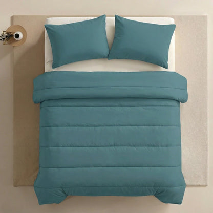 Ein ordentlich gemachtes Bett mit Zelesta Comfort Uni Kissenbezügen Set von 2 in Seegrün (80x80cm) auf tealfarbener Mikrofaser-Bettwäsche, platziert auf einem hellen Flächenteppich neben einem kleinen runden Beistelltisch mit einer Vase und einem einzelnen Zweig.