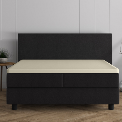 Ein modernes schwarzes Polsterbett mit einem cremefarbenen Doppel Jersey Topper Spannbettlaken aus gestrickter Baumwolle auf einem Holzboden vor einer hellgrau getäfelten Wand, gepaart mit einer kleinen Pflanze auf einem hölzernen Beistelltisch.