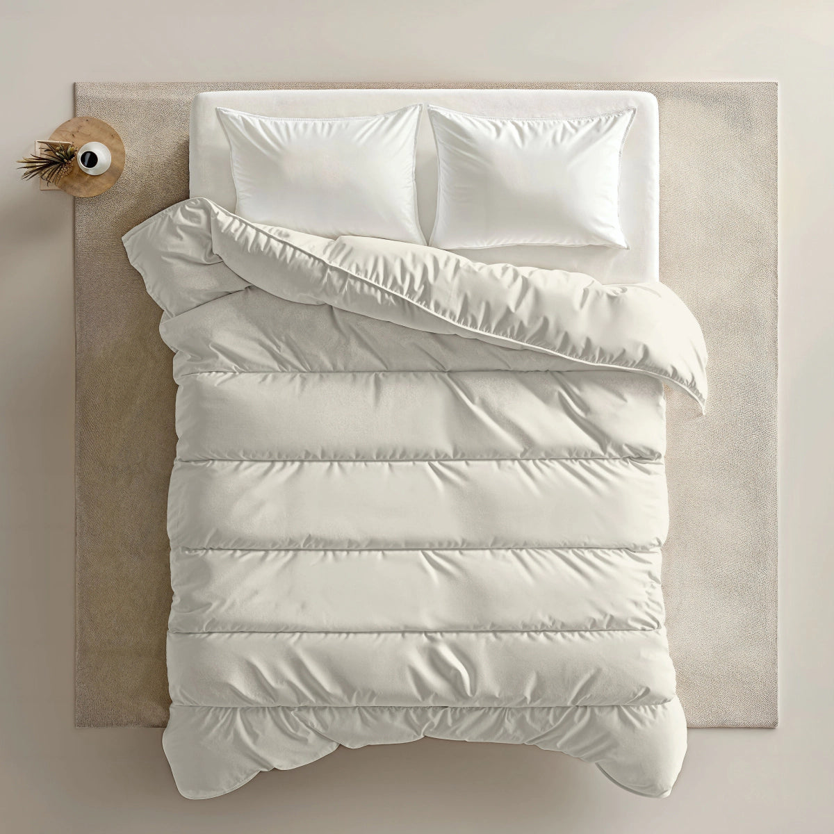 Ein ordentlich gemachtes Bett mit weißen Kissen und einer Zelesta Everyday Uni Bettdecke - Creme, von oben gesehen neben einem kleinen runden Nachttisch mit einer Vase und einer getrockneten Pflanze auf einem beigen Teppich.