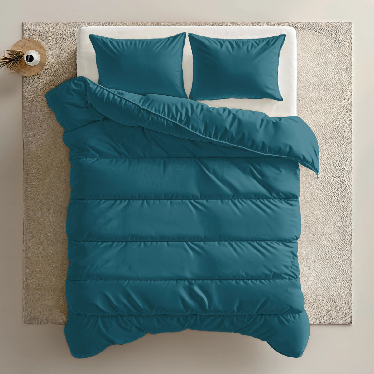 Ein ordentlich gemachtes Bett mit tealfarbener Bettwäsche, zwei passenden Kissen und einer Zelesta Everyday Uni Bettdecke - Nachtblau, die teilweise umgeschlagen ist, steht auf einem hellen Teppich mit einer kleinen Vase auf dem Nachttisch.