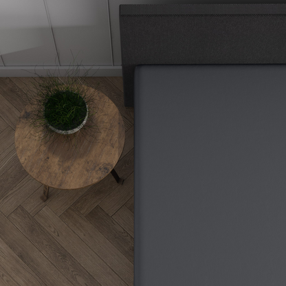 Ein runder Beistelltisch aus Holz mit einer Topfpflanze steht neben einem Bett aus dunkelgrauen Doppel Jersey Topper Spannbettlaken, das auf einem Holzboden mit Fischgrätenmuster steht und eine gemütliche Dreamhouse-Atmosphäre verbreitet.