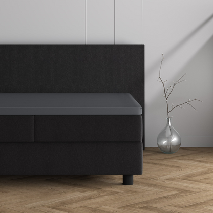 Ein modernes, minimalistisches schwarzes Bett mit einem dunklen Kopfteil zeigt den Doppel Jersey Topper Spannbettlaken - Grau neben einer klaren Glasvase mit dünnen Zweigen auf einem hellen Holzboden mit Fischgrätenmuster vor einer schlichten Wand.