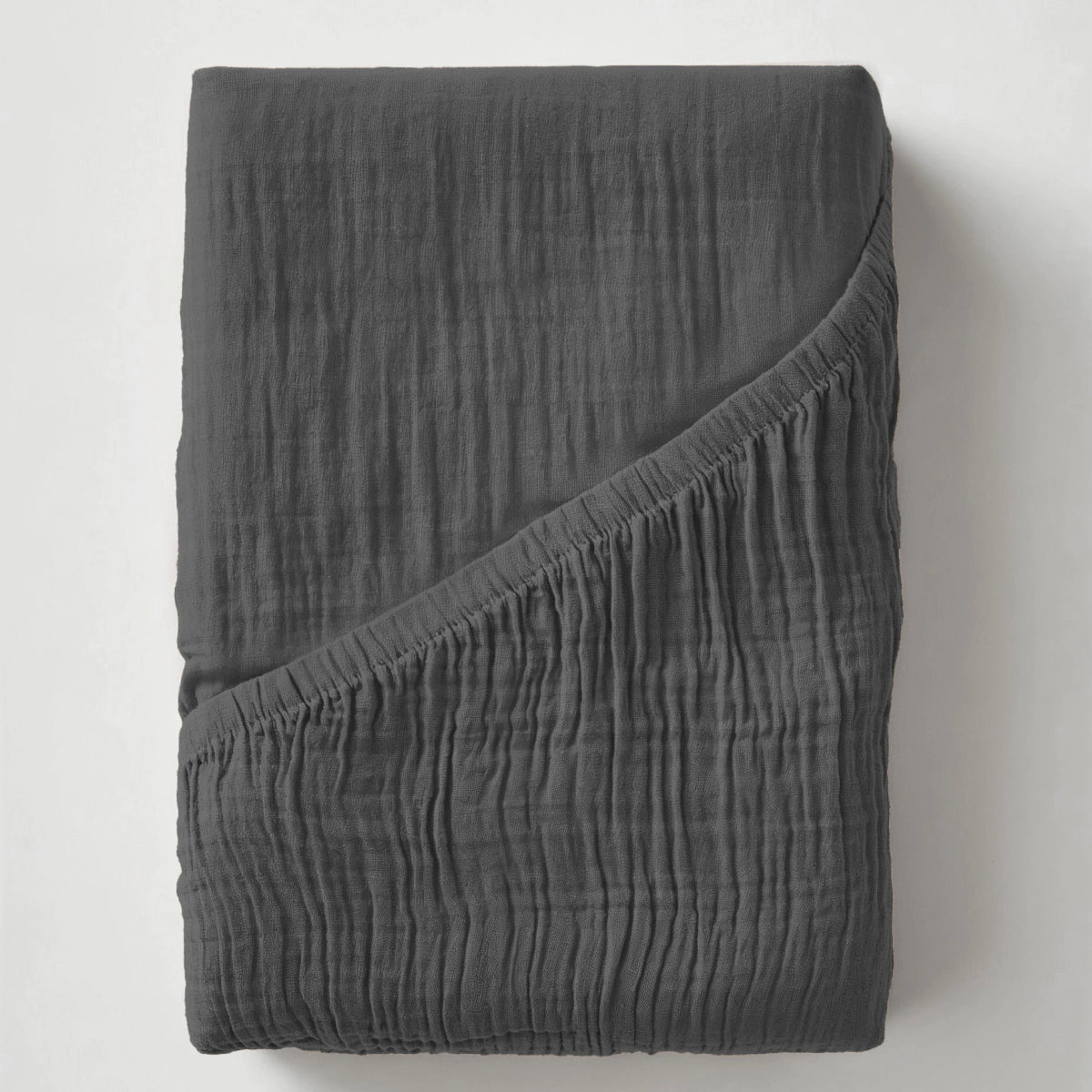 Ein dunkelgraues Musselin Spannbettlaken - Anthrazit, das mit einer Ecke nach unten gefaltet wird, um seine knittrige Struktur zu enthüllen, erinnert an einen gemütlichen, modernen, japanisch inspirierten Schlafzimmerstil.