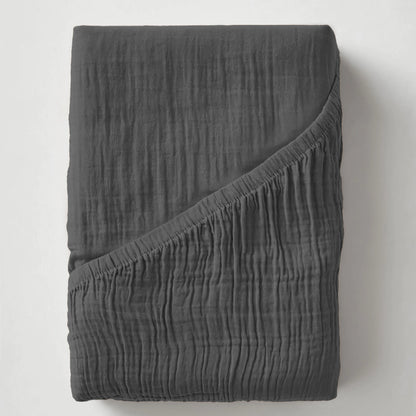 Ein dunkelgraues Musselin Spannbettlaken - Anthrazit, das mit einer Ecke nach unten gefaltet wird, um seine knittrige Struktur zu enthüllen, erinnert an einen gemütlichen, modernen, japanisch inspirierten Schlafzimmerstil.