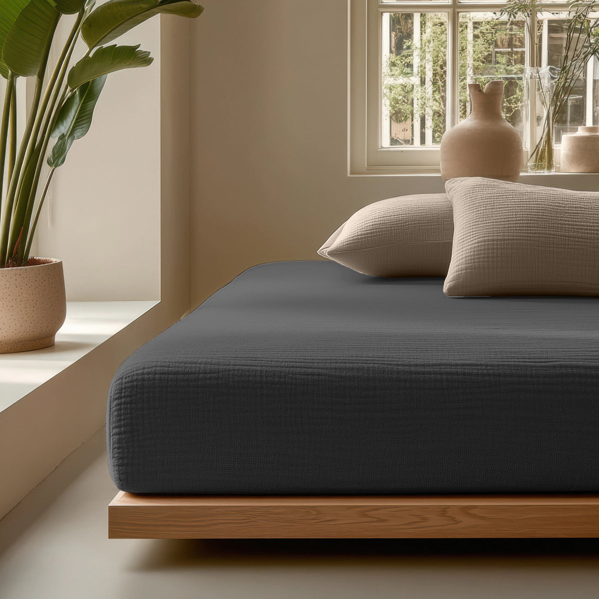 Ein minimalistisches Schlafzimmer mit einem Holzpritschenbett aus Musselin Spannbettlaken - Anthrazit, beigen Kissen, Topfpflanzen und Keramikvasen am Fenster mit Blick auf üppiges Grün.