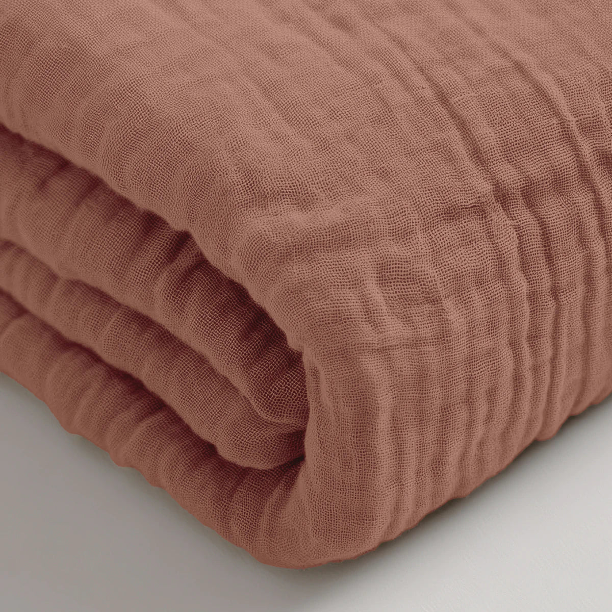 Ein gefaltetes Musselin Spannbettlaken - Dusty-Rose mit einer strukturierten Oberfläche ist in Nahaufnahme auf einer weißen Fläche zu sehen.