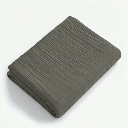 Ein sauber gefaltetes Musselin Spannbettlaken in Graugrün mit strukturierter Oberfläche und minimalistischem Japandi-Stil wird auf einer schlichten weißen Fläche präsentiert.