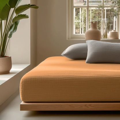 Ein modernes Schlafzimmer mit einem niedrigen Holzbettgestell, einer orangefarbenen Tagesdecke, grauen Kissen, Musselin Spannbettlaken - Taupe, einer großen Topfpflanze und einem Fenster, das natürliches Licht hereinlässt.