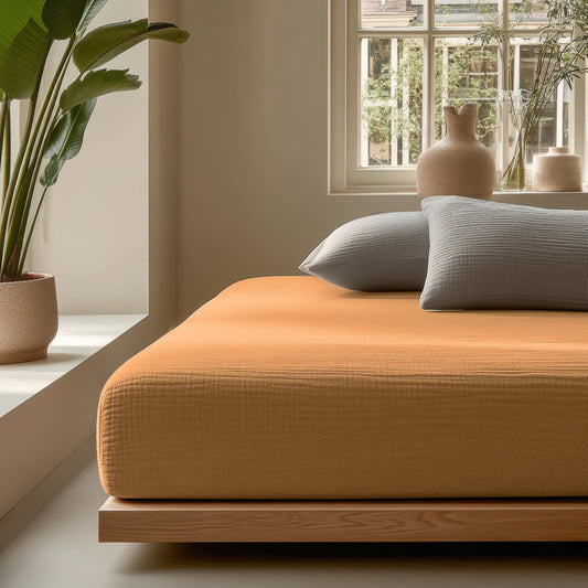 Ein modernes Schlafzimmer mit einem niedrigen Holzbettgestell, einer orangefarbenen Tagesdecke, grauen Kissen, Musselin Spannbettlaken - Taupe, einer großen Topfpflanze und einem Fenster, das natürliches Licht hereinlässt.
