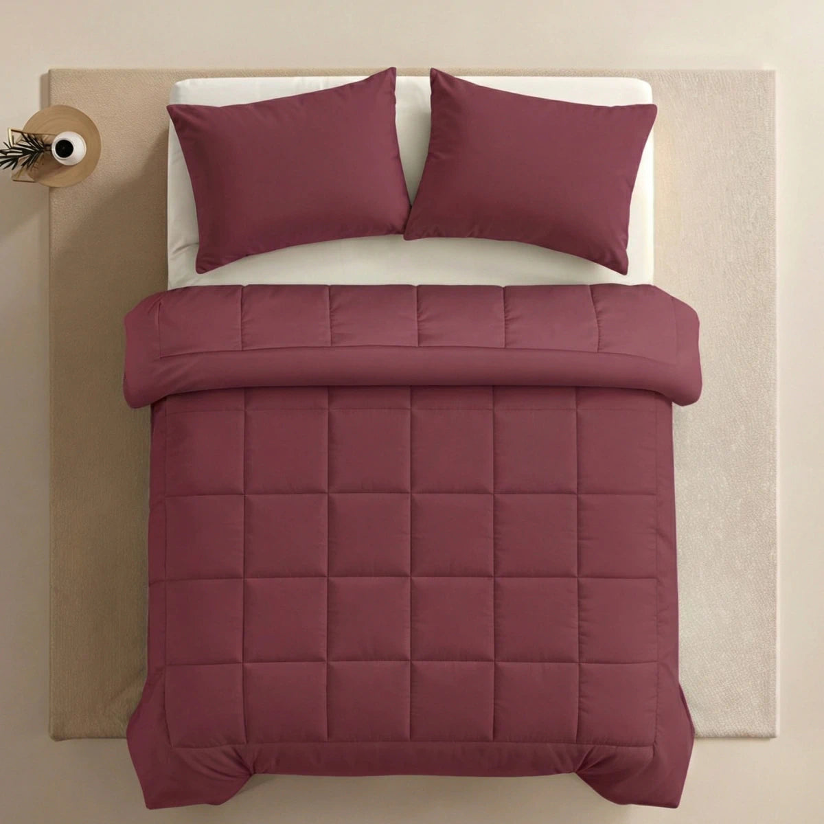 Ein ordentlich gemachtes Bett mit der Zelesta Pure Uni Bettdecke in Bordeauxrot und zwei passenden Kissen, auf einem beigen Teppich mit einem kleinen runden Nachttisch mit einer Vase.