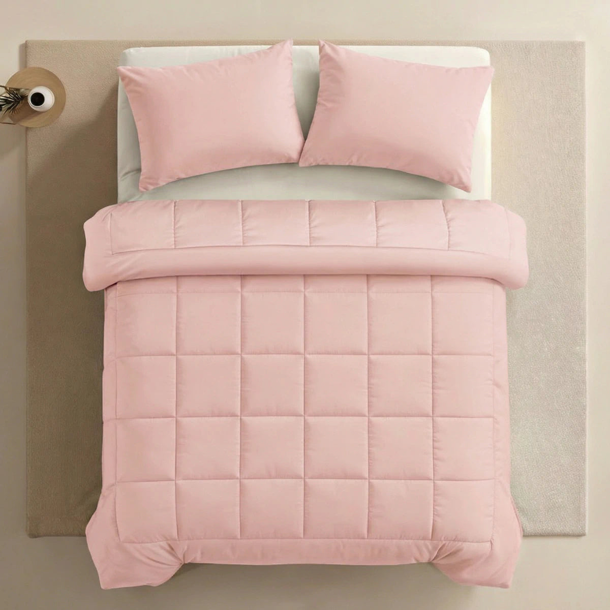 Ein ordentlich gemachtes Bett mit der Zelesta Pure Uni Bettdecke - Zartrosa, einer hellrosa Steppdecke mit zwei passenden Kissen auf einer beigen Oberfläche, betont den ganzjährigen Komfort und den gemütlichen Stil.