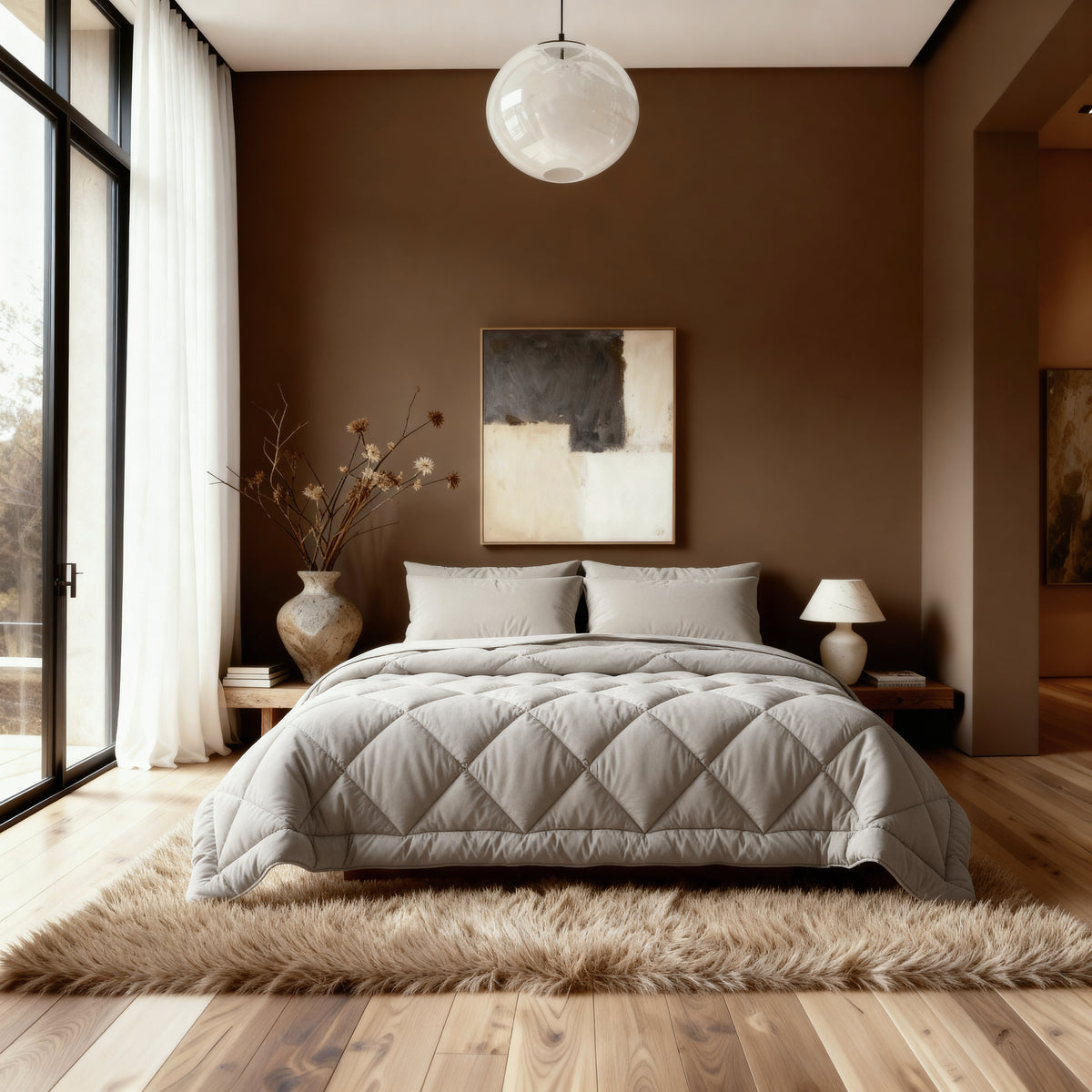 Ein modernes Schlafzimmer mit einem ordentlich gemachten Bett mit dem Zelesta Royalbed Diamant - Sand-Beige, beiger Bettwäsche, einer großen Vase mit getrockneten Zweigen, abstrakter Wandkunst, einer Pendelleuchte und bodentiefen Fenstern mit transparenten Vorhängen.